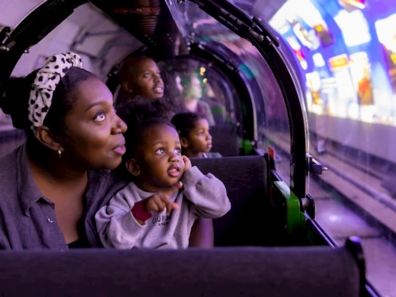 A mother and child sitting in the carriage of a miniature train. They are looking out the window at a video being projected onto the walls of the train platform.