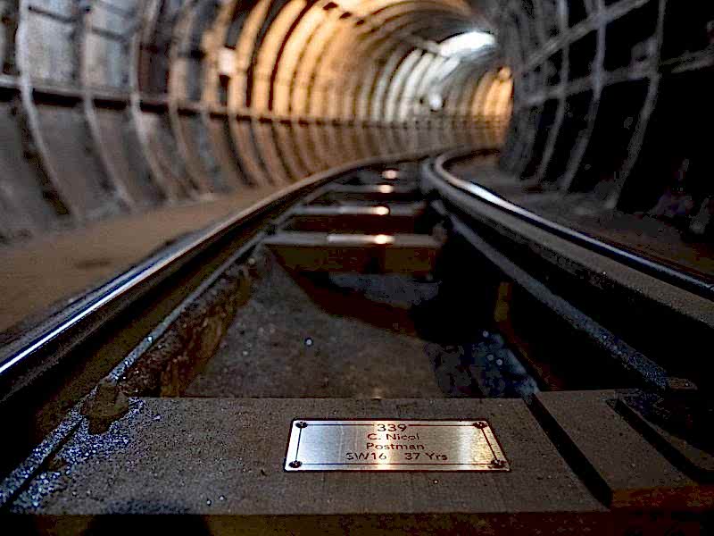 A closeup view of a silver coloured metal plaque, fixed to a wooden sleeper rail on the floor of the Mail Rail tunnel.