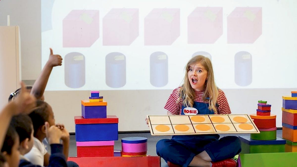 A learning facilitator is sitting on the floor surrounded by children. She holds a game and looks at them with a surprise face.
