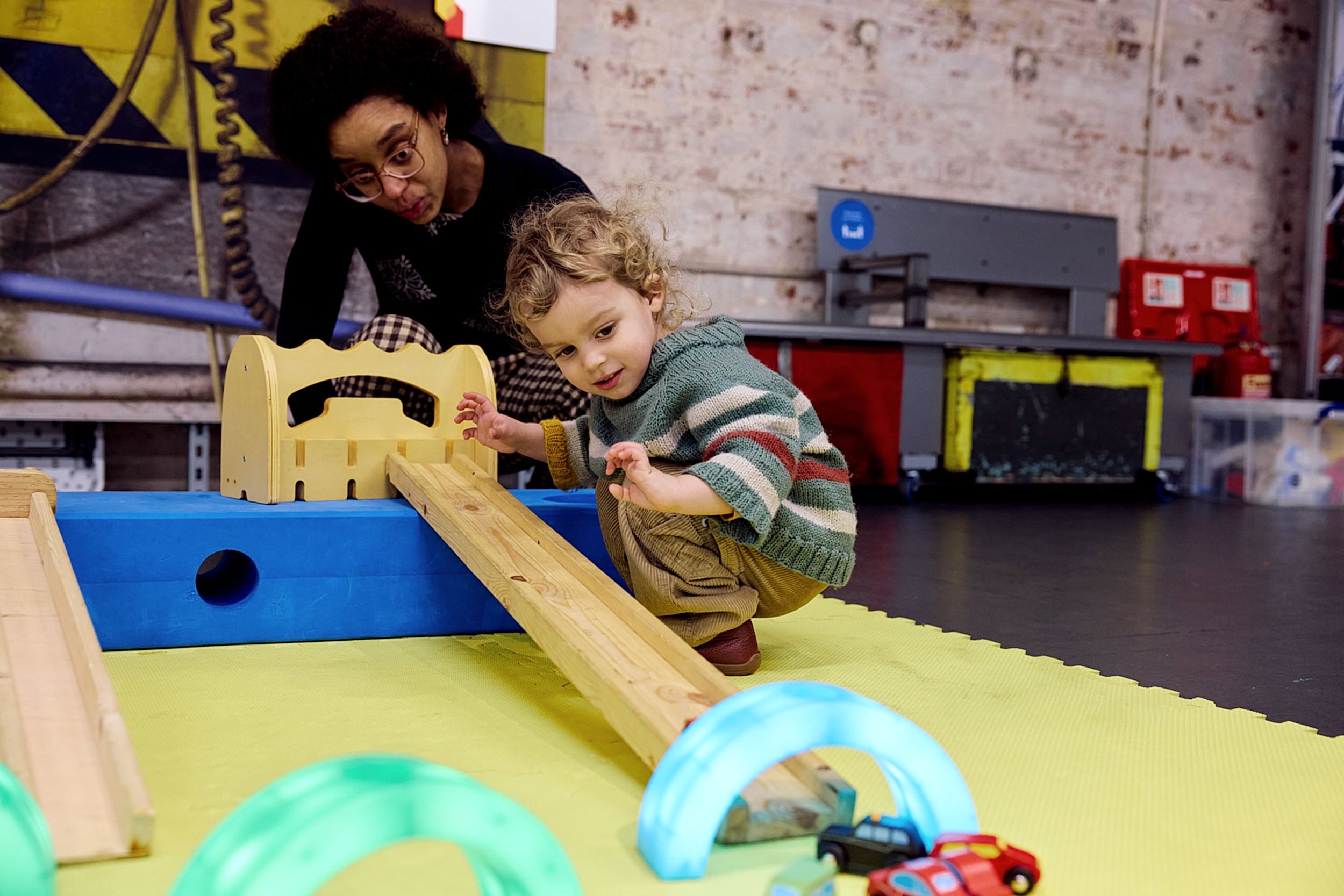A little boy crouches by a ramp and watches his little toy car speed down it, through a colourful tunnel.